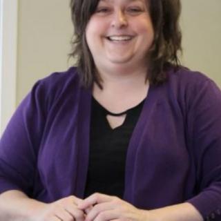 A woman is smiling while sitting indoors, wearing a purple sweater.