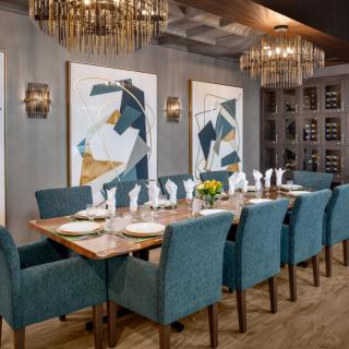 An elegantly decorated dining room featuring a long table with blue chairs, modern artwork on the walls, and sophisticated chandeliers.