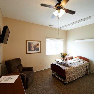 A cozy bedroom featuring a single bed, armchair, and wall-mounted TV, illuminated by natural light from the window.