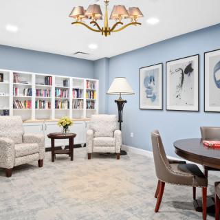 A cozy living room with bookshelves, armchairs, and a round table, featuring elegant artwork on the walls.