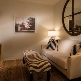 A cozy living room with a white sofa, decorative pillows, a bench with a zigzag pattern, a lamp on the side table, and wall art illuminated by warm lighting.