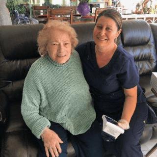 An elderly woman sitting on a couch with her caregiver, both smiling.