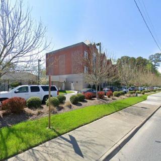 A street view featuring a brick building, lined with trees and parked cars along the sidewalk.