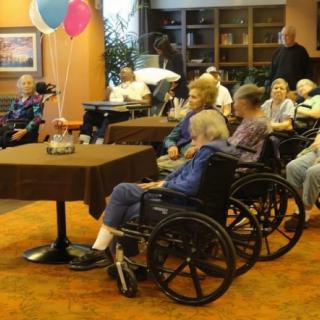 A group of elderly people in wheelchairs gathered in a nursing home setting with balloons and decorations.