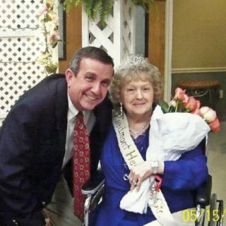 An elderly woman wearing a tiara and sash sits in a wheelchair holding flowers, accompanied by a smiling man.