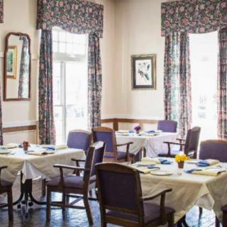 A cozy dining room with neatly arranged tables and chairs, adorned with table settings and surrounded by windows with patterned curtains.