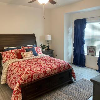 A cozy bedroom featuring a bed with red floral bedding and decorative pillows, a bedside table with a lamp, and a window with blue curtains.