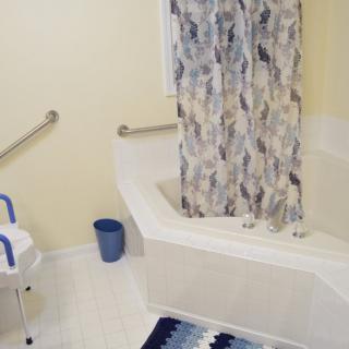 An accessible bathroom featuring a bathtub with grab bars, a shower curtain, a safety chair, and a small trash bin on the floor.