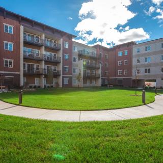 A residential apartment complex with a green courtyard and balconies under a partly cloudy sky.