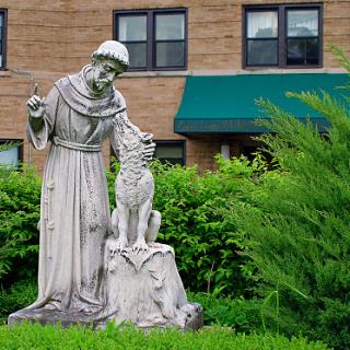 A statue of a saint interacting with a dog, surrounded by lush greenery and set against a brick building.