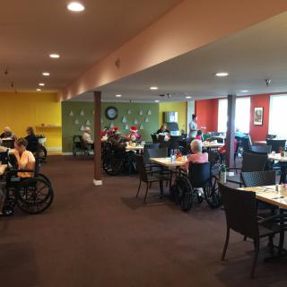 A dining area in a nursing home, with residents seated in wheelchairs, enjoying their meals.