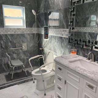 A modern, accessible bathroom featuring marble tiles, a walk-in shower with grab bars, and a toilet equipped with safety rails.