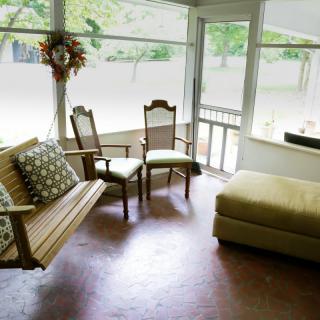A cozy porch with a wooden swing, chairs, and an ottoman bathed in natural light.