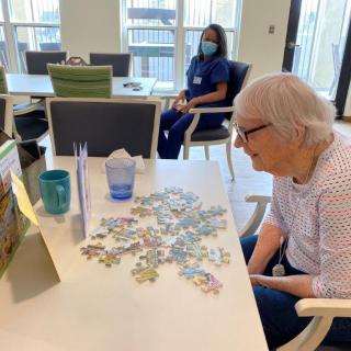 An elderly person engages in a puzzle activity at a nursing home, with a caregiver in the background.