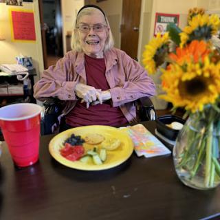 A cheerful elderly woman enjoying a colorful plate of fresh fruit, surrounded by sunflowers and a red cup.
