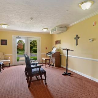 A small chapel interior with chairs, a cross on the wall, and an organ near the window.