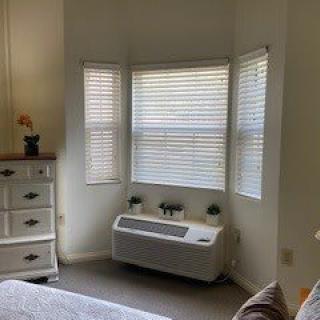 A cozy bedroom corner featuring a bay window with closed blinds, a dresser with an ornament, and an air conditioning unit beneath the window.
