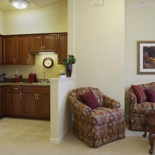 A cozy living room leading to a small kitchen area, featuring comfortable armchairs and wooden cabinets.