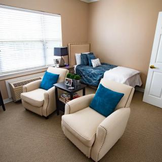 A cozy bedroom with a neatly made bed and two comfortable chairs, complemented by soft lighting from a large window.