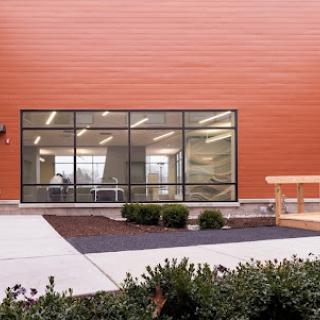 A modern building featuring large windows and an orange exterior, with well-maintained landscaping in front.