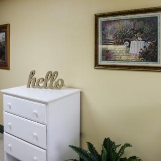 A cozy interior featuring a white chest of drawers with decorative paintings on the wall and a welcoming 'hello' sign.