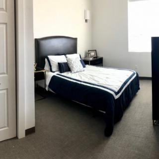 A neatly arranged bedroom featuring a bed with dark bedding, nightstands on either side, and natural light coming through a window.