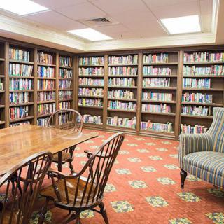 A cozy library reading room with wooden bookshelves filled with books, a large table, chairs, and a comfortable armchair on a patterned carpet.