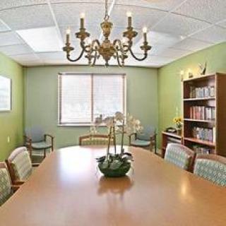 A well-lit conference room featuring a large table, chairs, a chandelier, and a bookshelf against green walls.