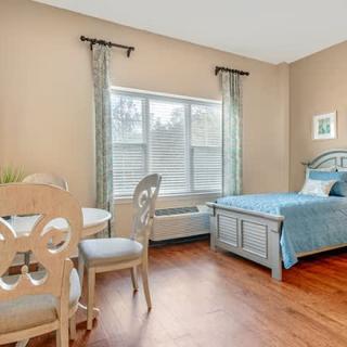 A cozy bedroom with a single bed, a small dining table and chairs, a window with sheer curtains, and hardwood flooring.