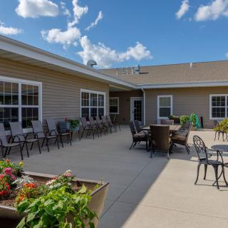 A sunny patio area with outdoor seating and flower pots, set against a residential building under a blue sky with clouds.