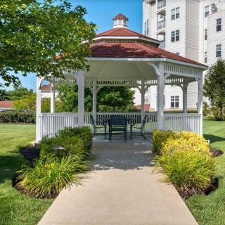 A picturesque gazebo surrounded by greenery and a pathway, set against the backdrop of a tall building.