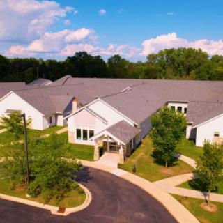 An aerial view of a residential building surrounded by lush greenery and a winding driveway under a partly cloudy sky.
