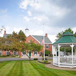 A scenic view of a gazebo on a well-manicured lawn with a brick building and trees in the background under a cloudy sky.