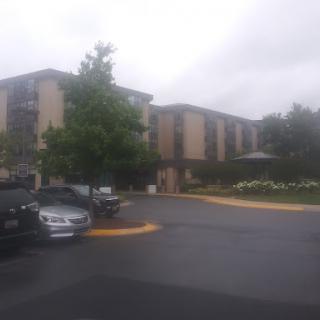 A hotel building seen on a cloudy day, featuring a parking lot and greenery.