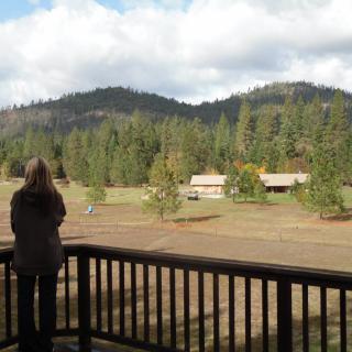 A person stands on a deck overlooking a scenic landscape with cabins, trees, and mountains.