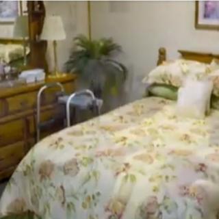 A cozy bedroom with floral bedding, a wooden dresser with a mirror, an indoor plant, and a walker beside the bed.