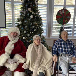 A festive indoor setting with Santa Claus and two elderly individuals sitting by a decorated Christmas tree.