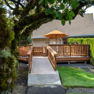 A serene outdoor deck with a ramp, shaded by an umbrella, surrounded by lush greenery.