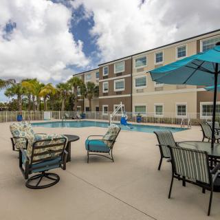 A view of an outdoor swimming pool with a seating area and umbrellas, situated in front of a multi-story apartment building under a cloudy sky.