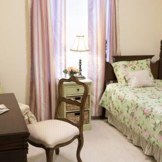 A cozy bedroom with vintage furniture, featuring a single bed with floral bedding, a nightstand with a lamp, and elegant window curtains.