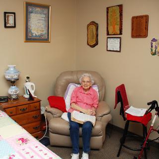 An elderly person sitting comfortably in an armchair, surrounded by personal items and wall decorations.