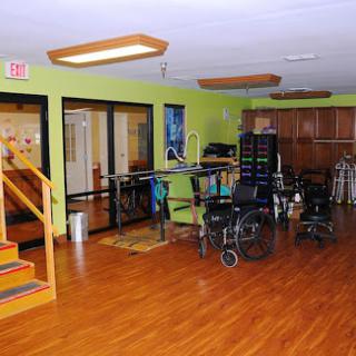 A spacious rehabilitation room equipped with wheelchairs and therapy equipment, featuring vibrant green walls and a warm wooden floor.