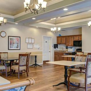 A well-lit dining area with tables and chairs, adjacent to a kitchen with wooden cabinets and chandeliers overhead.