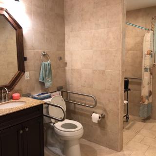 An accessible bathroom with tiled walls and flooring, featuring a toilet with handrails, a sink with a mirror above, and a shower area.