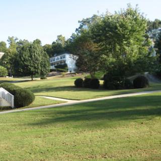 A peaceful park landscape with grassy areas, trees, pathways, and white buildings in the background.