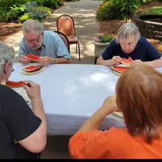 A group of people enjoying watermelon outdoors at a table in a park setting.