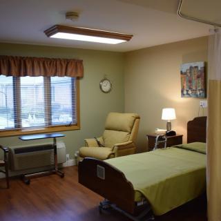 A well-maintained hospital room with a single patient bed, comfortable chair, and natural light from the window.