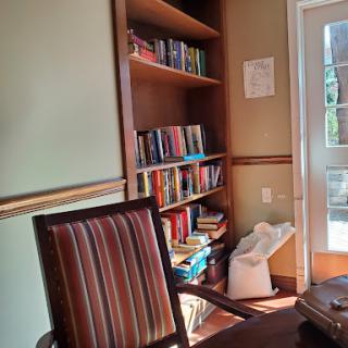 A cozy office space featuring a bookshelf filled with books, a comfortable chair, and natural light coming through the door.