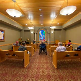 A serene chapel interior with wooden benches and a stained glass window, where several people are seated in contemplation.