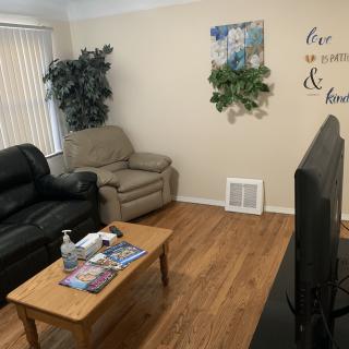 A cozy living room with a sofa, armchair, and a television. A coffee table is placed in the center, adorned with magazines and hand sanitizer. The walls feature decorative elements such as wall art and an indoor plant.